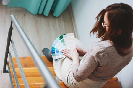 Young woman designer with long red hair holding color samples sitting on the stairs at home