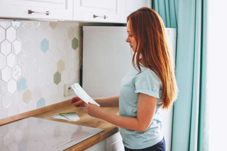 Young woman designer with long red hair holding color samples in the bright kitchen at home