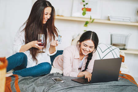 Young carefree brunette girls friends in casual with glass of wine buy online by credit card on the laptop, shopping from homeの写真素材