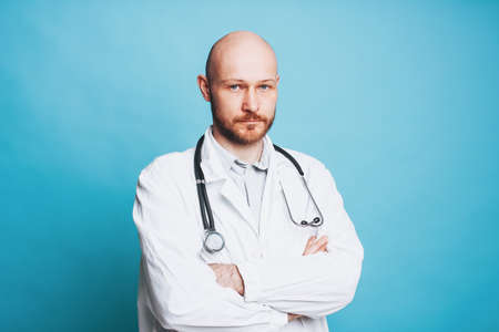 Attractive serious man bald bearded doctor looking at camera isolated on the blue background