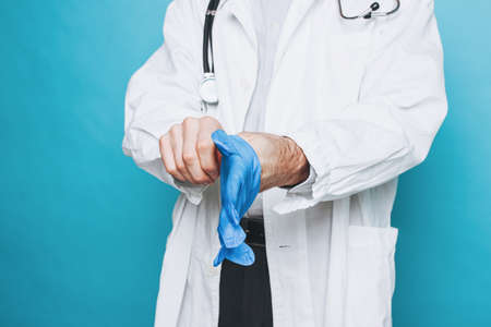 Crop photo of man doctor in white coat puts on rubber medical gloves  isolated on the blue backgroundの写真素材