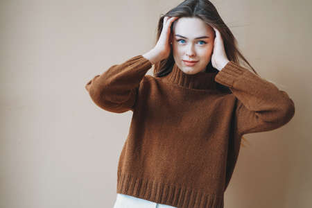 Young beautiful long brown-haired hair girl with blue eyes in brown knitted sweater looking at camera on the beige backgroundの写真素材