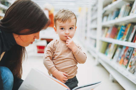 Cute baby boy toddler child in bookstore with mother with open bookの写真素材