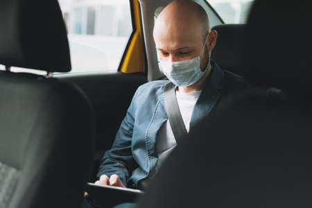 Bald man businessman in medical face mask using tablet mobile phone inside yellow car taxi. Concept of coronavirus quarantineの写真素材