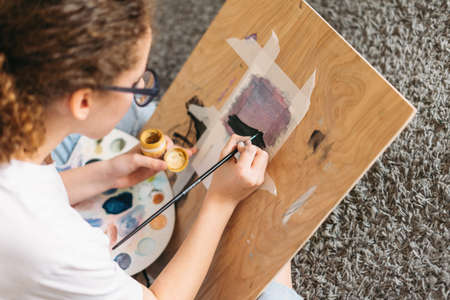 Curly hair girl teenager in glasses in white t-shirt sitting on floor painting on wooden desk at the homeの写真素材