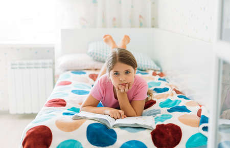 Cute tween girl in pink t-shirt reading book lying on bed in bright room at the homeの写真素材