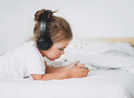 Cute toddler girl with fair hair in headphones with mobile on bed in bright interiorの写真素材