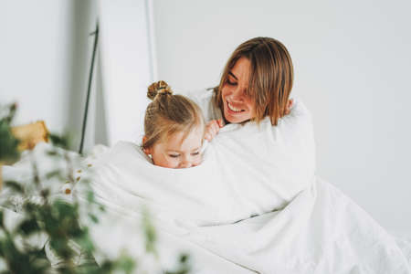Cute toddler girl long fair hair big grey eyes looking at camera having fun with mother on bed stay at homeの写真素材