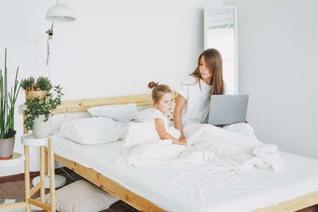Strict mother freelancer with little daughter want to work on laptop in bed in bright interior at homeの写真素材