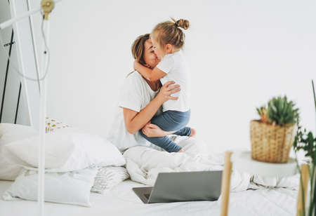 Cute toddler girl with long fair hair having fun with mother on bed in bright interiorの写真素材