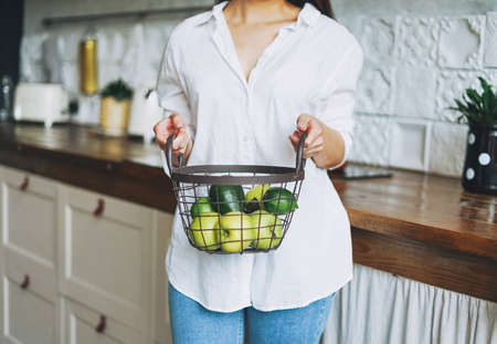 Crop photo of young woman in white shirt hold basket with green fruits and vegetables in hands on kitchenの写真素材