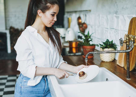Young woman washes dishes with wooden brush with natural bristles at window in kitchen. Zero waste conceptの写真素材
