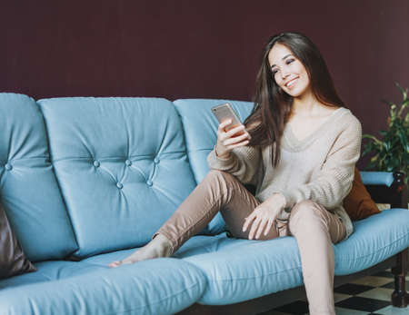 Smiling brunette asian young woman in casual using mobile sitting on sofaの写真素材