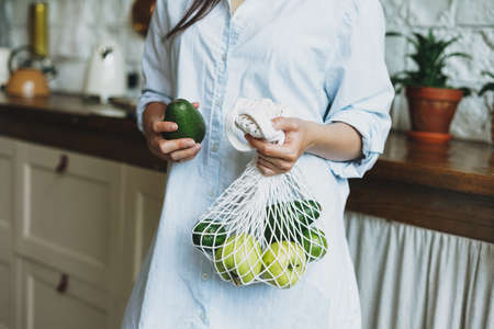 Young woman in shirt hold knitted rag bag shop with avocado and green apples in hands on kitchenの写真素材