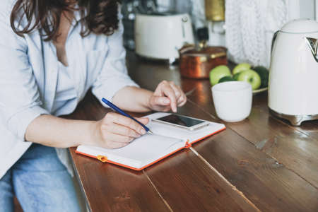 Brunette woman in casual doing notes in daily book with and using mobile on kitchen at homeの写真素材