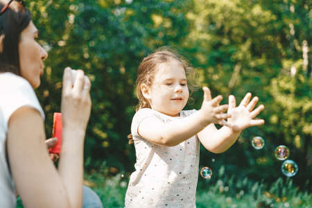 Cute toddler girl blows on soap bubbles with his mother in sunny summer dayの写真素材