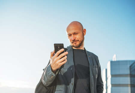 Handsome adult bald bearded man businessman in suit using mobile phone on background of blue sky and...