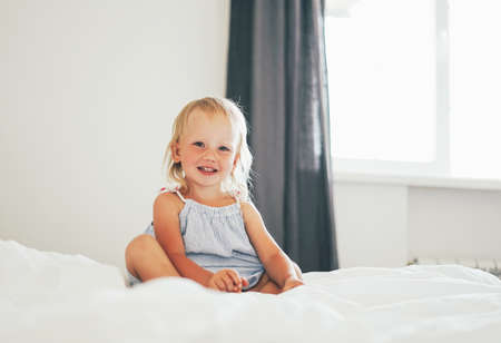 Cute blonde baby girl on bed with white bed linen at bright interiorの写真素材
