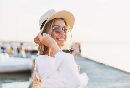 Close up portrait of beautiful blonde young woman in white shirt and straw hat on sunset