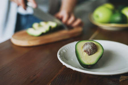 Female hands cut avocado on wooden working surface in kitchen at homeの写真素材