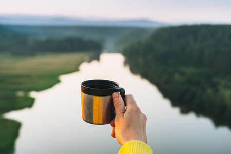 Female hand with metal mug on background of beautiful view on morning calm river, forest and hills. Local tourism conceptの写真素材