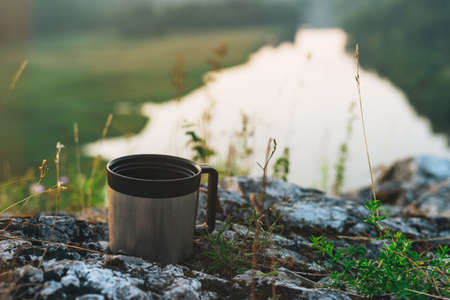 Metal mug on background of beautiful view on morning calm river, forest and hills. Local tourism conceptの写真素材