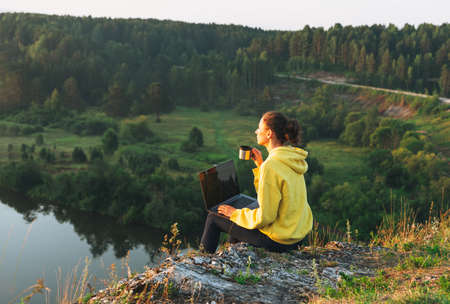 Young woman girl traveler freelancer in yellow hoodie with opened laptop looking at sunrise on beautiful view backgroundの写真素材