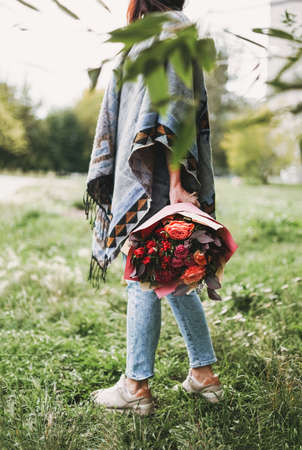 Young brunette woman in poncho and jeans holds beautiful bouquet of different flowers in red tonesの写真素材