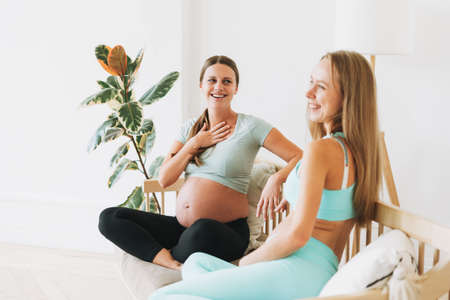 Two friends pregnant women in sports uniforms laughing sitting on the couch in bright interiorの写真素材