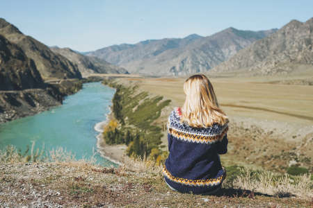 Thinking young blonde woman in blue dress sitting on stones on background of mountainsの写真素材