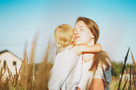 Young woman mom with baby girl on hands hug in backyard of cottage in sunset lightの写真素材