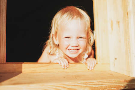 Portrait of cute blonde baby girl in window of wooden house, country side