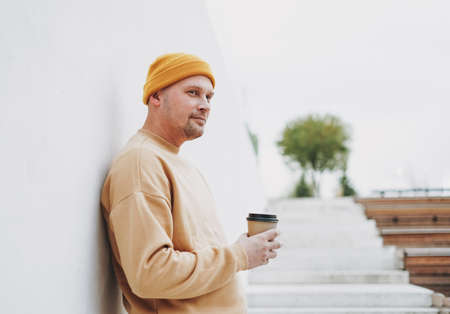 Portrait of stylish man hipster in yellow hat with paper cup of coffee at city