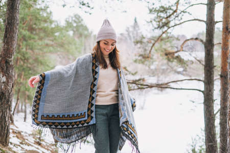 Young brunette beautiful woman in hat and gray poncho in the winter forest