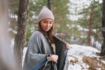 Young brunette beautiful woman in hat and gray poncho in the winter forest