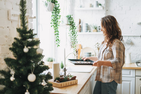 Attractive smiling woman with curly hair in plaid shirt bakes cookies at bright kitchen at homeの写真素材