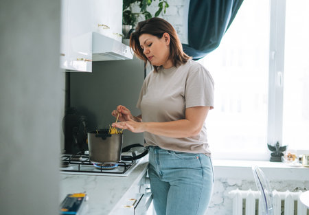 Beautiful smiling brunette young woman plus size body positive using laptop in kitchen at homeの写真素材