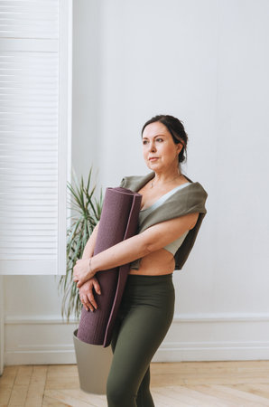 Attractive middle aged brunette woman in sportswear with yoga mat in the light studioの写真素材