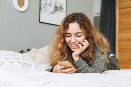 Beautiful smiling teenager girl with curly hair using mobile phone sitting on the window sill, remote communicationの写真素材