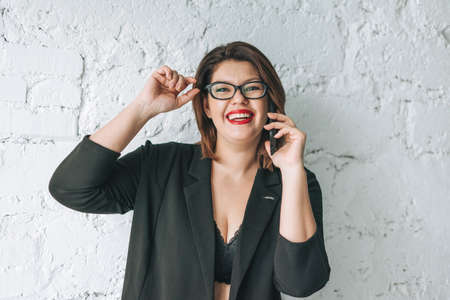 Beautiful smiling young woman business lady in glasses and black jacket with cellphone against the white wallの写真素材