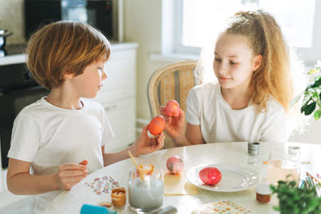 Two siblings brother and sister toddler boy tween girl painting easter eggs on kitchen at home on the spring sunny dayの写真素材