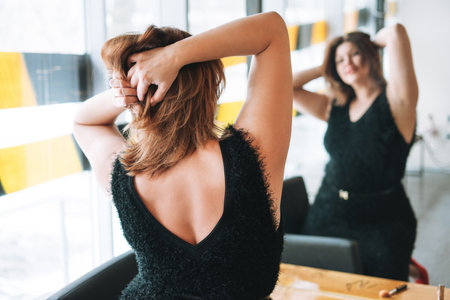 Beautiful smiling young woman plus size body positive in black evening dress hair style with red lipstick in hands looking at mirror in the beauty studioの写真素材
