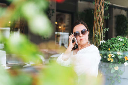 Beautiful brunette middle aged woman in sunglasses in white clothes talking on mobile phone in hand with coffee at terrace of the summer cafeの写真素材