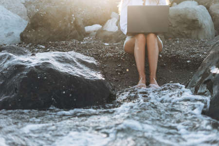 Crop photo of young carefree beautiful freelance businesswoman in white shirt working at laptop on seashore on sunriseの写真素材