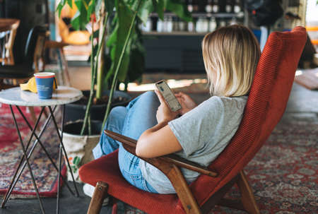 Young blonde woman in casual clothes using social media on mobile phone sitting on the chair at cafe, people from behindの写真素材