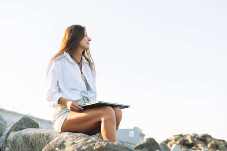 Young carefree beautiful freelance businesswoman with long hair in a white shirt working at laptop on seashore on the sunriseの写真素材