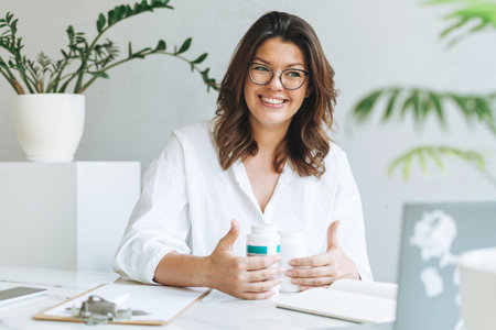 Young smiling brunette woman nutritionist plus size in white shirt working at laptop on table with house plant in the bright modern office. Doctor communicates with the patient onlineの写真素材