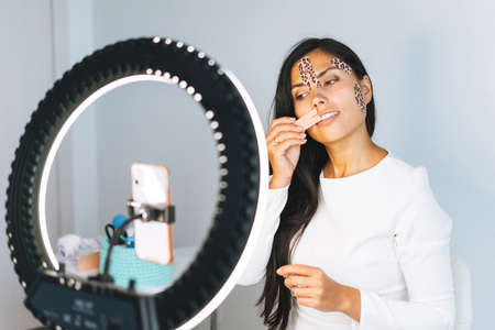 Young beautiful brunette woman with long hair cosmetologist makes face taping looks in the mirror in...