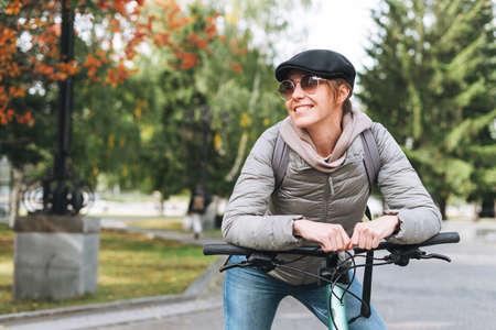 Portrait of fashionable young pretty woman in cap and sunglasses on bicycle with thermo mug on sunny autumn day in cityの写真素材