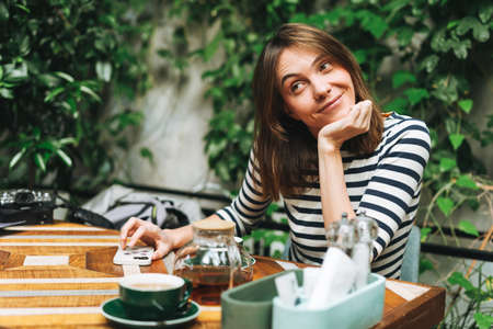 Young smiling brunette woman in casual clothes using social media on mobile phone sitting at green cafeの写真素材